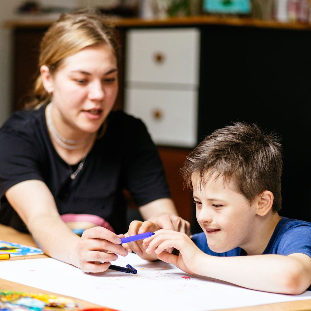Close up of young female teacher sitting at desk with a Down syndrome schoolboy. Color painting on the paper for disabled kids, autism childs who are down syndrome and student teacher. Close up of young female teacher sitting at desk with a Down syndrome schoolboy. Color painting on the paper for disabled kids, autism childs who are down syndrome and student teacher.